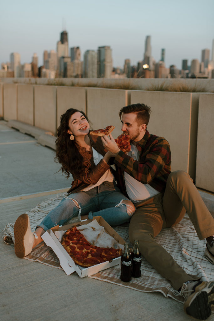 boyfriend and girlfriend feed each other pizza on a chicago parking garage rooftop picnic with the chicago skyline in the background