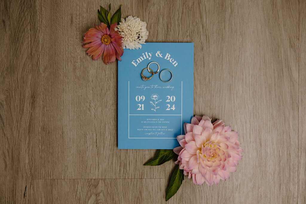 Simple blue wedding invitation with pink flowers and gold and silver wedding rings sitting on light wood floors.  