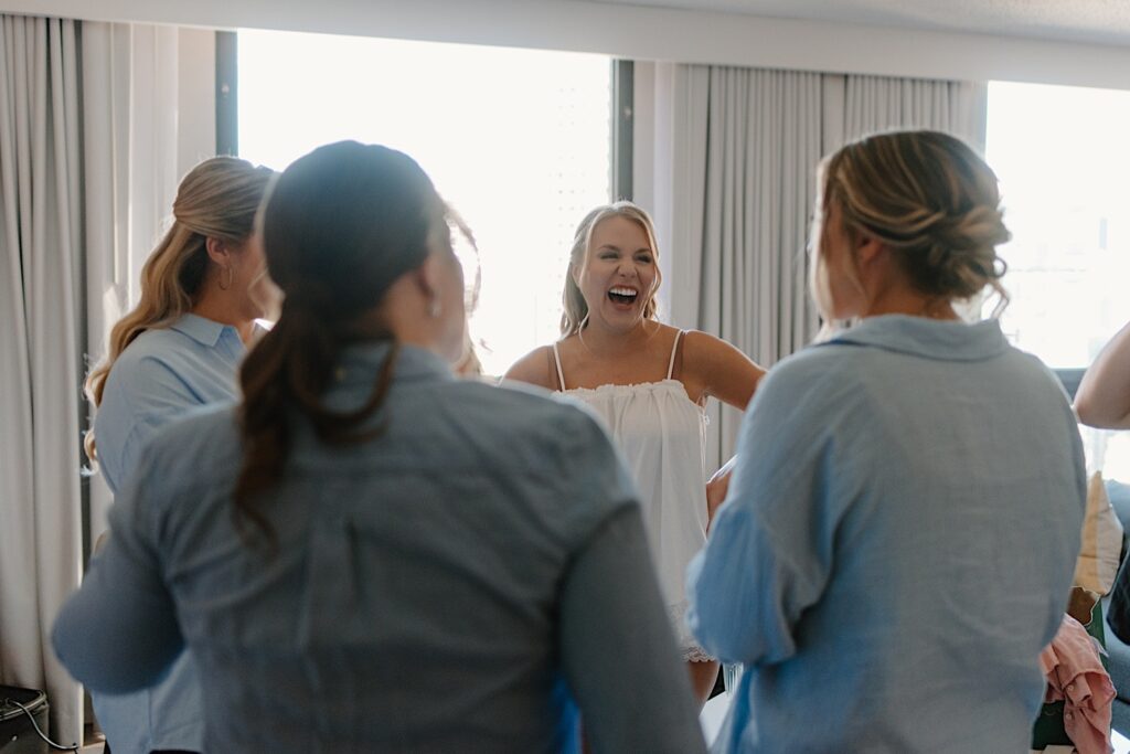 A bride stands with her bridesmaids laughing the morning of her wedding.  She's wearing a white linen pajama set and her bridesmaids are wearing oversized blue button ups.  They're getting ready for the wedding day in a Chicago hotel.