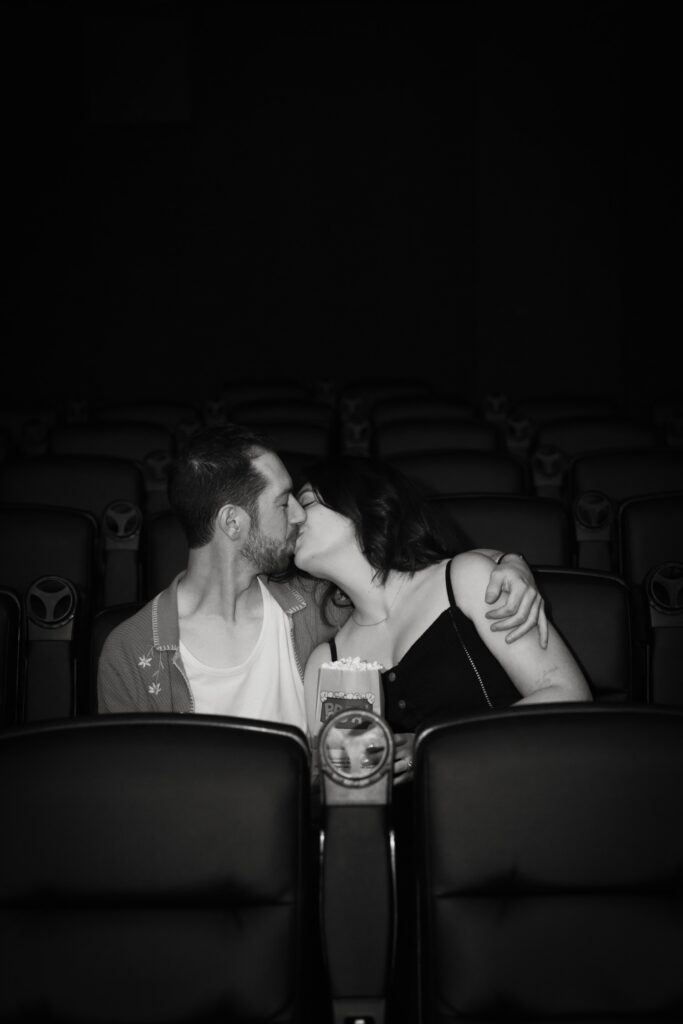 A couple kisses during their engagement session at a movie theater in Logan Square in Chicago.  The fiancée is wearing a black dress and the fiancé is wearing a button up with embroidery on it. 