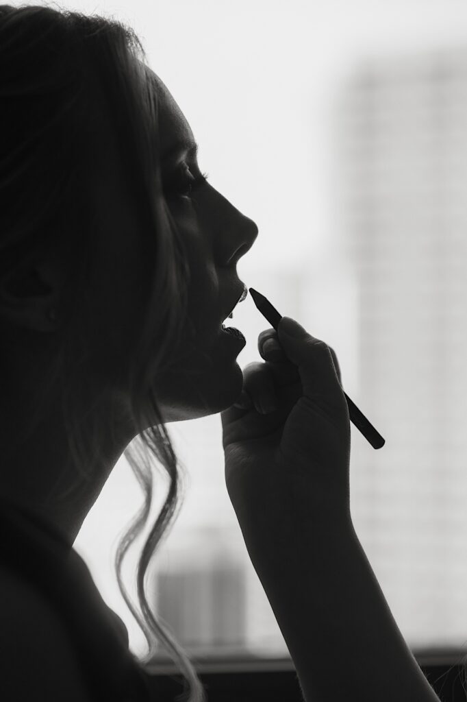 A bride gets lipliner put on her lips she's backlit by a large window overlooking the city of Chicago.