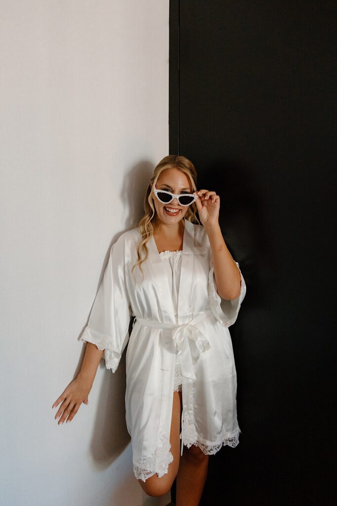 A bride stands in the corner of her hotel room with white and black walls, she's wearing a white linen pajama set and a silk wedding robe with white sunglasses in her Chicago hotel.