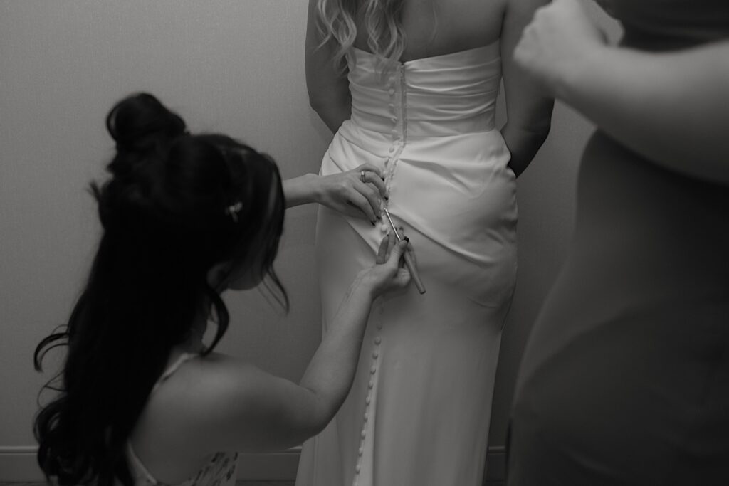 A bride stands with her back to the camera while a bridesmaid uses a crochet hook to button up her wedding dress. 