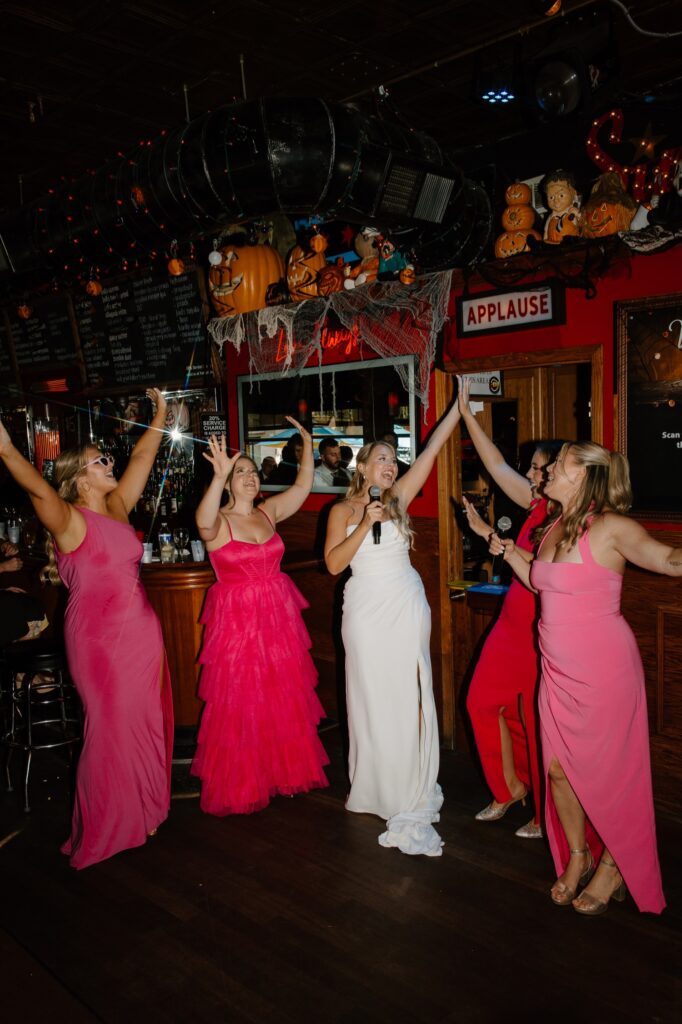 A bride sings with her bridesmaids at a Louie's pub in Chicago on her wedding day.  Her bridesmaids wear hot pink wedding dresses and the bride smiles at them as she sings.