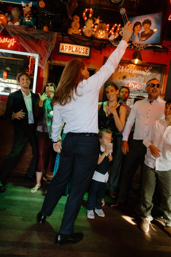 A groom celebrates his wedding day by singing karaoke at Louie's pub in Chicago.  The groom dresses casually in a white button up with navy blue pants.