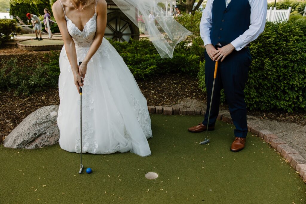 A bride and groom play mini golf on their wedding day in Chicago.  The bride is wearing a lace covered dress and the groom is wearing a blue vest and dress pants.