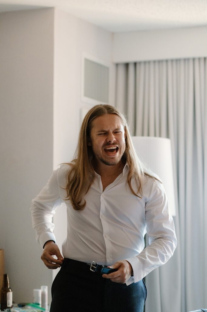 A groom puts on his belt the morning of his wedding getting ready with a sibling in his Chicago hotel room. 