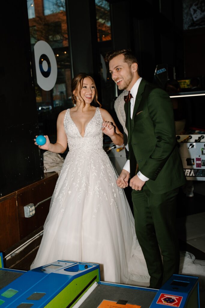 A bride and groom laugh and celebrate part of their wedding day at a Chicago arcade bar.  The bride holds a ski ball and laughs as her husband plays the game.  The bride is wearing an elegant dress with sparkling beads sewn to the bodice.  The groom is in a forest green suit. 