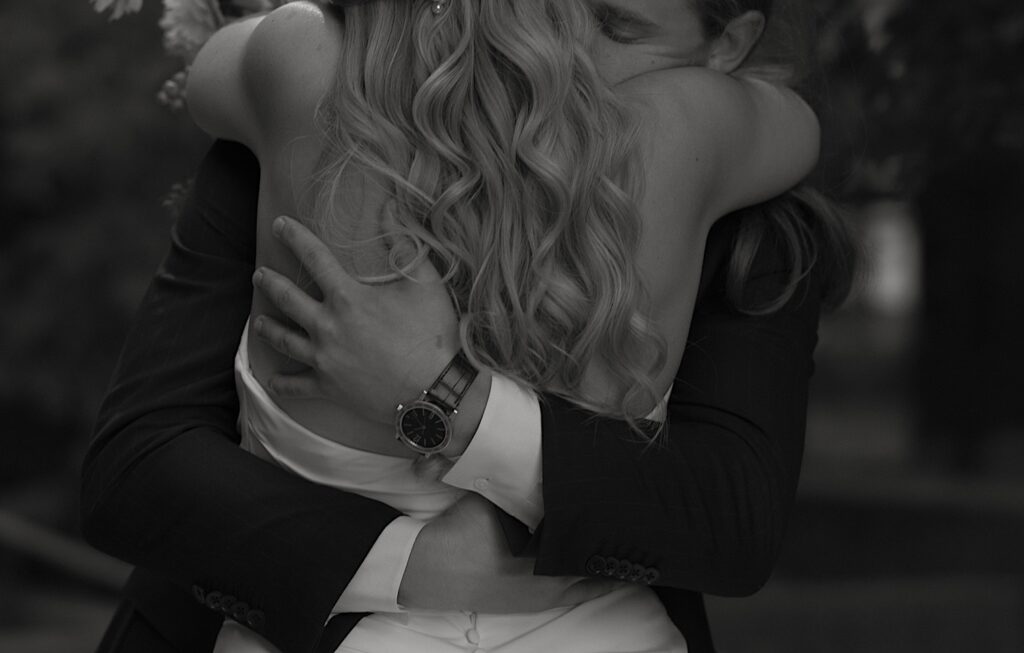 A close up black and white image of a groom hugging his bride the morning of their wedding you can see the brides loose curls and the grooms arms wrapped around her waist.