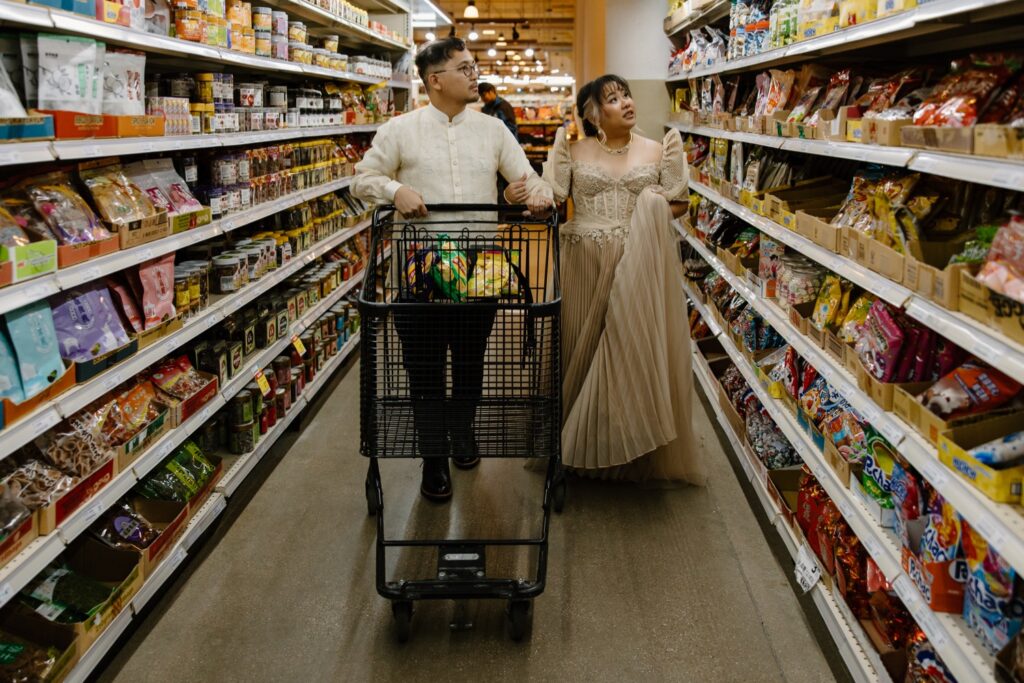 A bride and groom shop for some quick snacks on their wedding day from their favorite supermarket.  The aisles are lined with candy, the bride is wearing a beige wedding dress and the groom is wearing a traditional grooms attire for his wedding day.