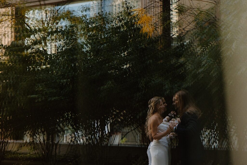 A reflection in a window of a skyscraper of a bride and groom sharing a moment together outside of their Chicago hotel room.  The bride and groom are out of focus, and the leaves are starting to change on the trees behind them for fall. 