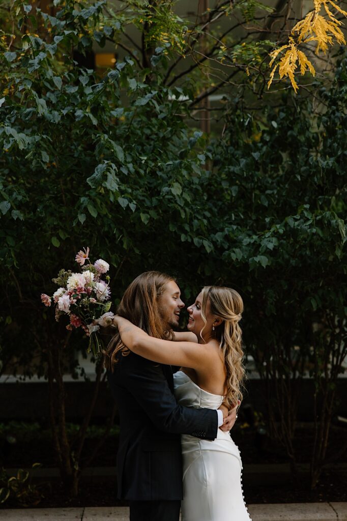 A bride and groom embrace the morning of their wedding.  The bride drapes her arms over her grooms shoulders holding a bouquet with pink and white wedding flowers.  Her dress is filled with pleats emulating a grecian style bodice. 