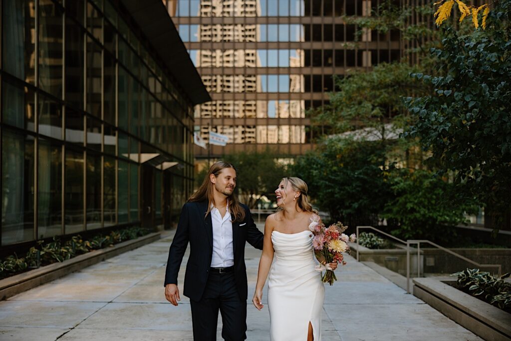 A bride and groom walk along side skyscrapers in the Loop in Chicago.  The bride laughs and the groom has his arm behind her back as they walk towards the camera. 