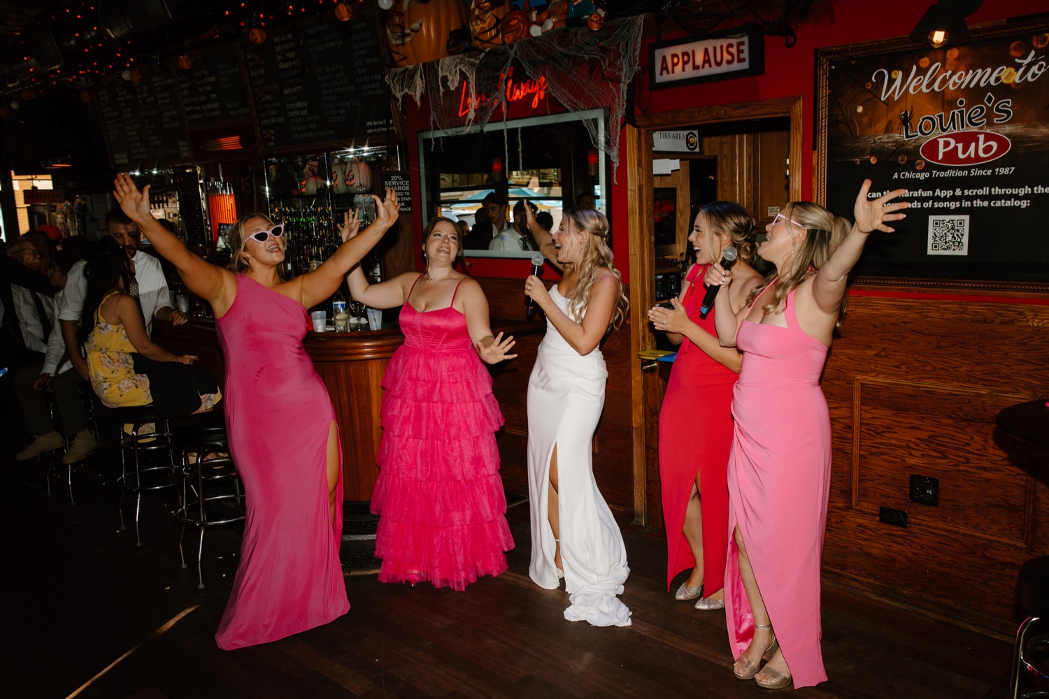 Bridesmaids in hot pink dresses sing and dance around their bride at a small pub in Chicago called Louie's Pub. The bride is wearing a wedding dress with grecian inspired pleats and a long train.