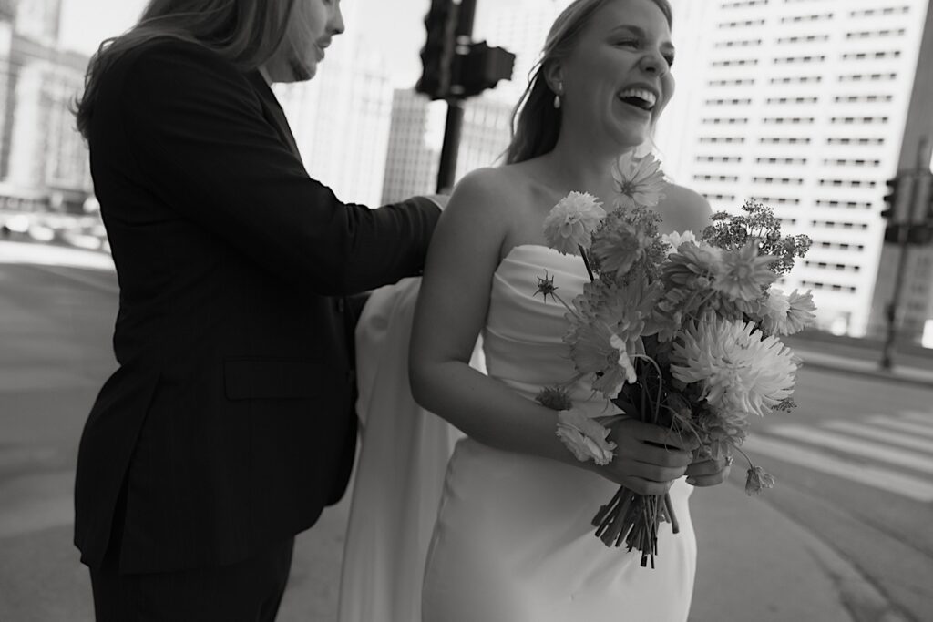 A bride and groom stand along Michigan Avenue by the River Walk in Chicago.  The bride laughs and the groom is holding the train of her grecian inspired wedding dress.  She holds a wedding bouquet filled with textured flowers like daisies and dahlias.