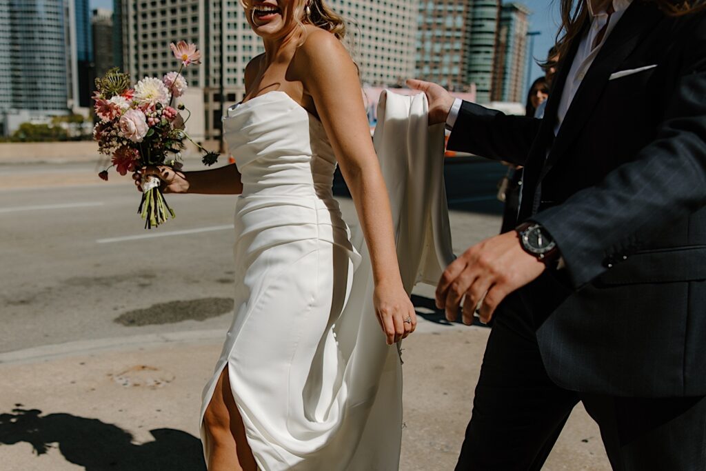 A bride and groom walk along Michigan Avenue by the River Walk in Chicago.  The bride laughs and the groom is holding the train of her grecian inspired wedding dress.  She holds a wedding bouquet filled with textured flowers like daisies and dahlias in shades of pink.