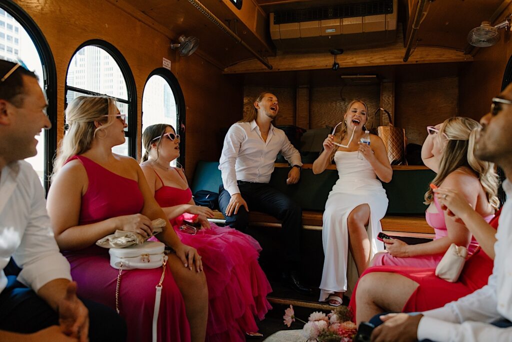 A bride and groom laugh with their wedding party on a Chicago wedding trolley.  The bridesmaids are wearing hot pink wedding attire and sunglasses. 