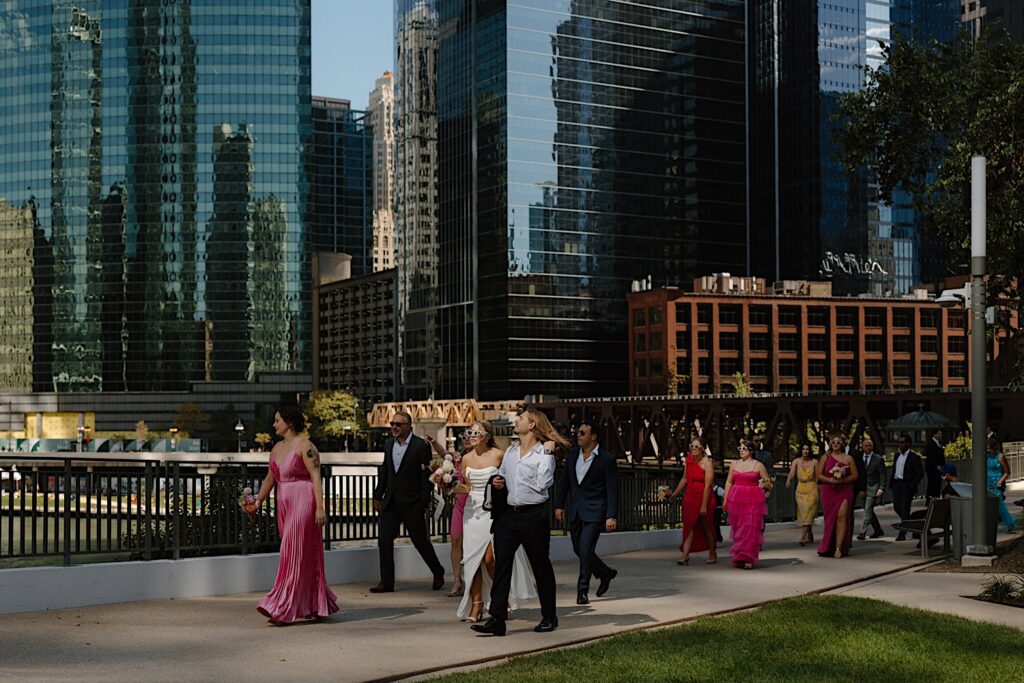 A groom and bride walk along the riverwalk in Chicago surrounded by skyscrapers to take wedding portraits on their wedding day.  The bridesmaids wear shades of pink and the groomsmen wear navy blue suits with casually buttoned white shirts. 