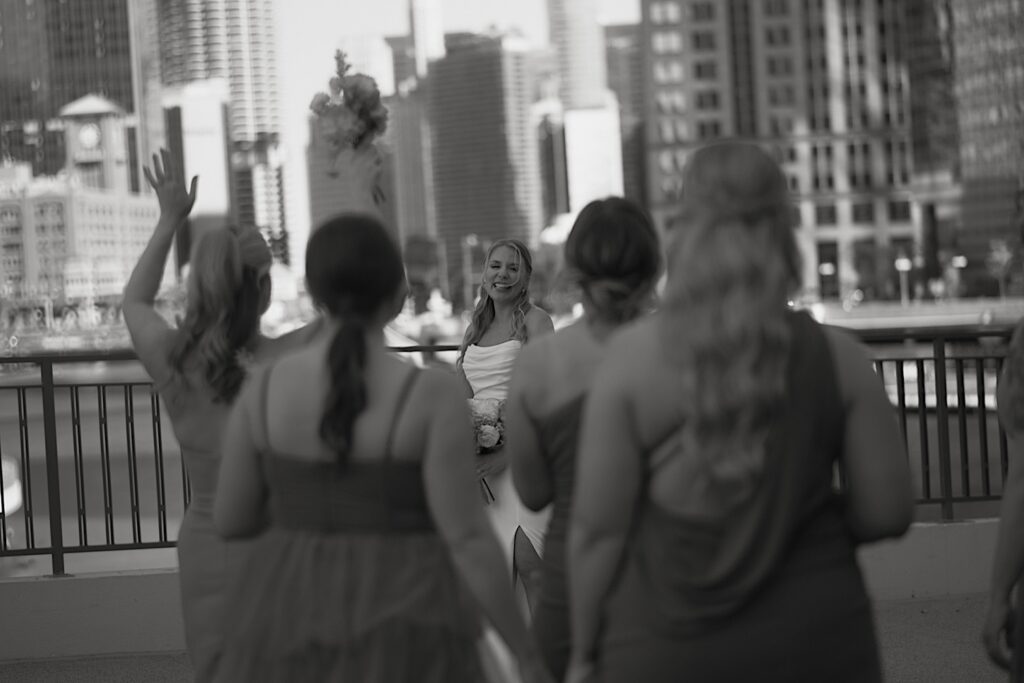 A bridesmaid tosses her wedding bouquet in the air and makes the bride laugh as all of the bridesmaids cheer on the bride during wedding portraits.  In the background of the bridal portraits you can see the Chicago river and many Chicago skyscrapers.