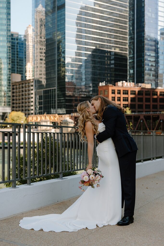 A bride and groom kiss while holding one another overlooking the Chicago River.  The bride is wearing a grecian inspired pleated dress with a train and is holding a wedding bouquet made up of flowers in shades of pink.