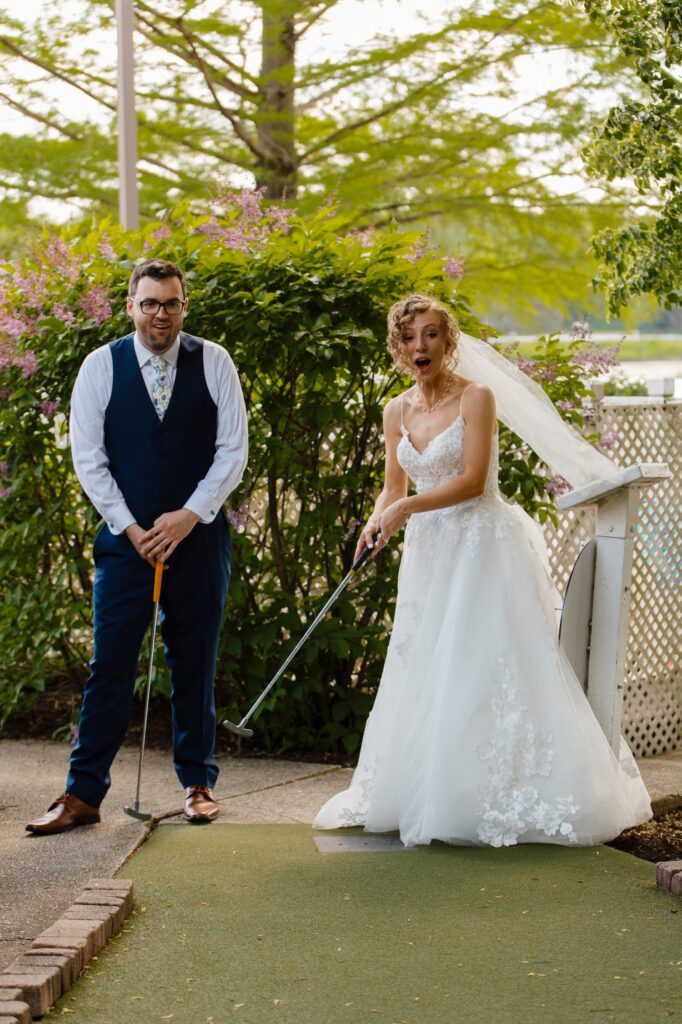 A bride and groom play mini golf in Chicago on their wedding day.  The groom look casual without his suit coat and the bride wears a dress covered in floral applique and a short veil while she plays. 