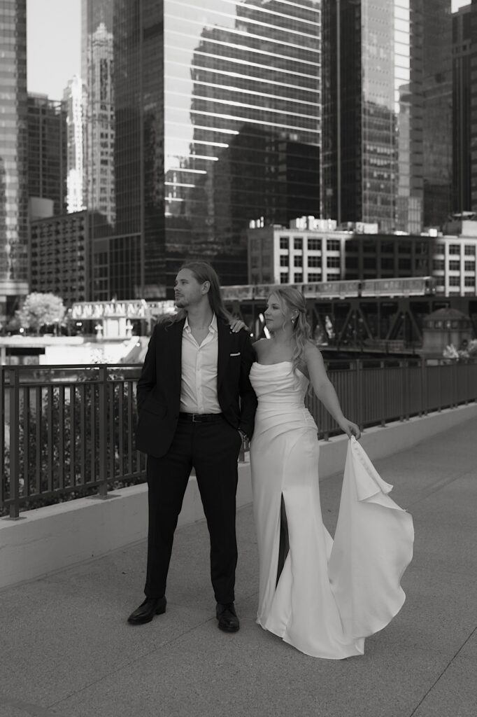 A bride and groom stand looking off into the distance.  The bride shows off her elegant pleated wedding dress by holding the train out to the side.  The groom is dressed casually for his Chicago wedding. 