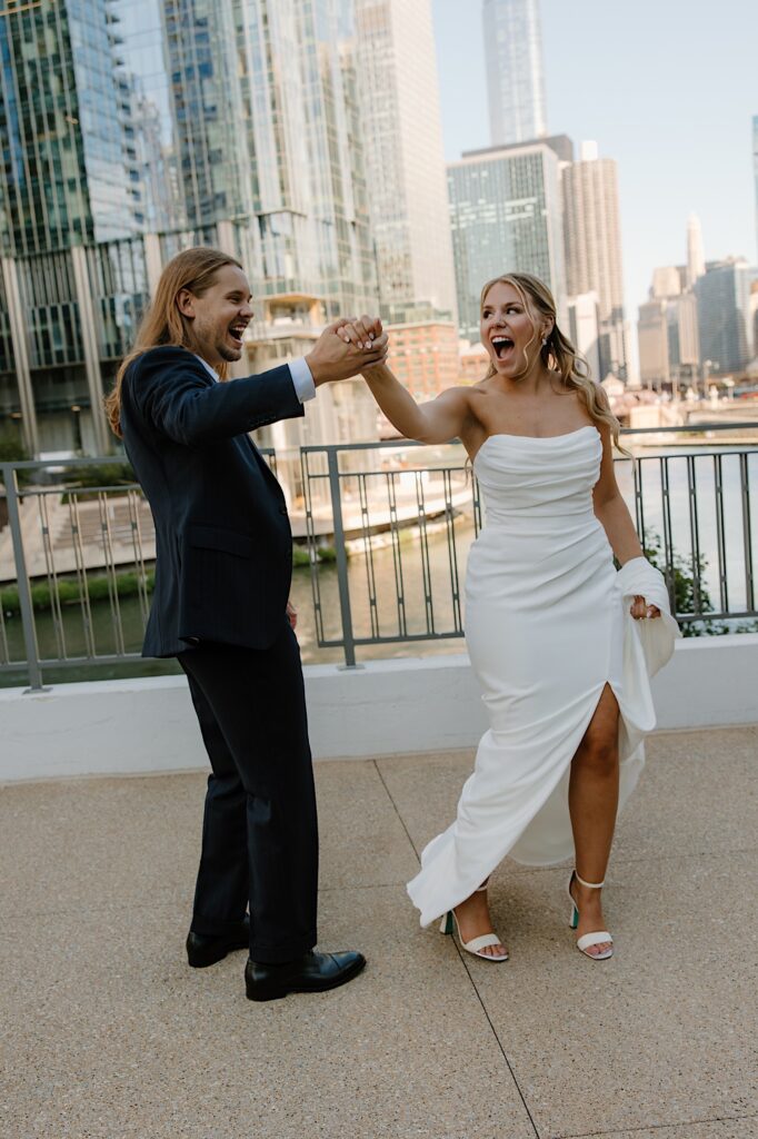 A bride and groom dance and laugh together during their wedding portraits overlooking the Chicago river.  The bride is wearing scrappy stiletto white heals and a grecian inspired pleated wedding gown.  The groom is wearing a navy blue suit. 