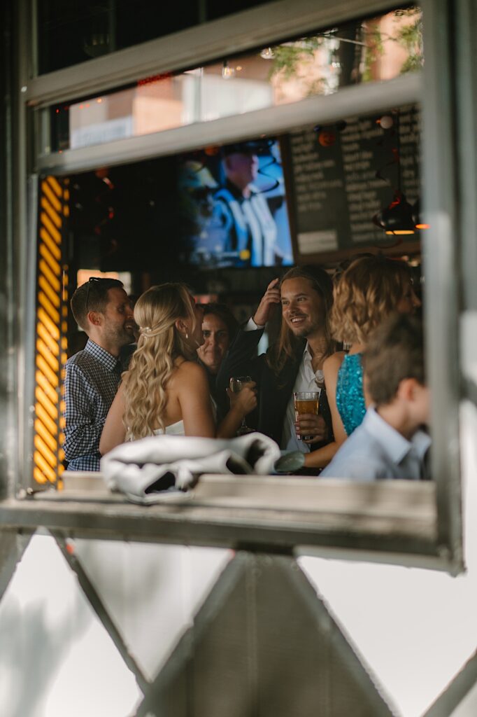 A photographer takes a photo through the window of a dive bar in Chicago of a bride and groom talking with guests before their wedding day. 