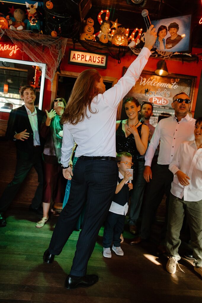 The groom stands while holding a microphone in his hand up in the air at Louie's Pub in Chicago on his wedding day. 