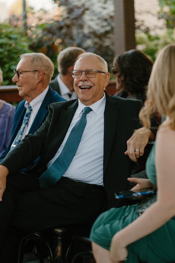 A guest in a black suit and green tie laughs with another guest before the outdoor rooftop wedding ceremony in Chicago.