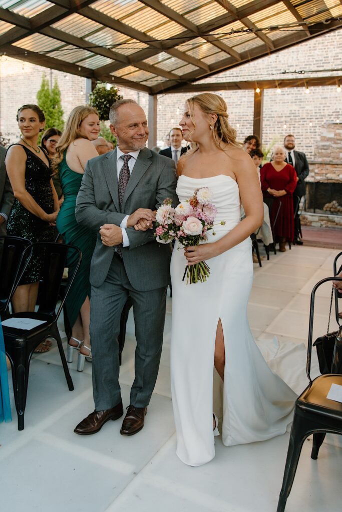 A bride and her father walk down the aisle to her groom on her wedding day.  She's holding a bouquet of pink dahlias and daisies for her fall wedding in Chicago. 