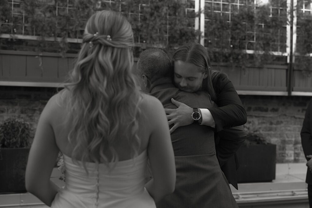 The groom hugs his brides father, you can see the back of the brides head in the foreground.