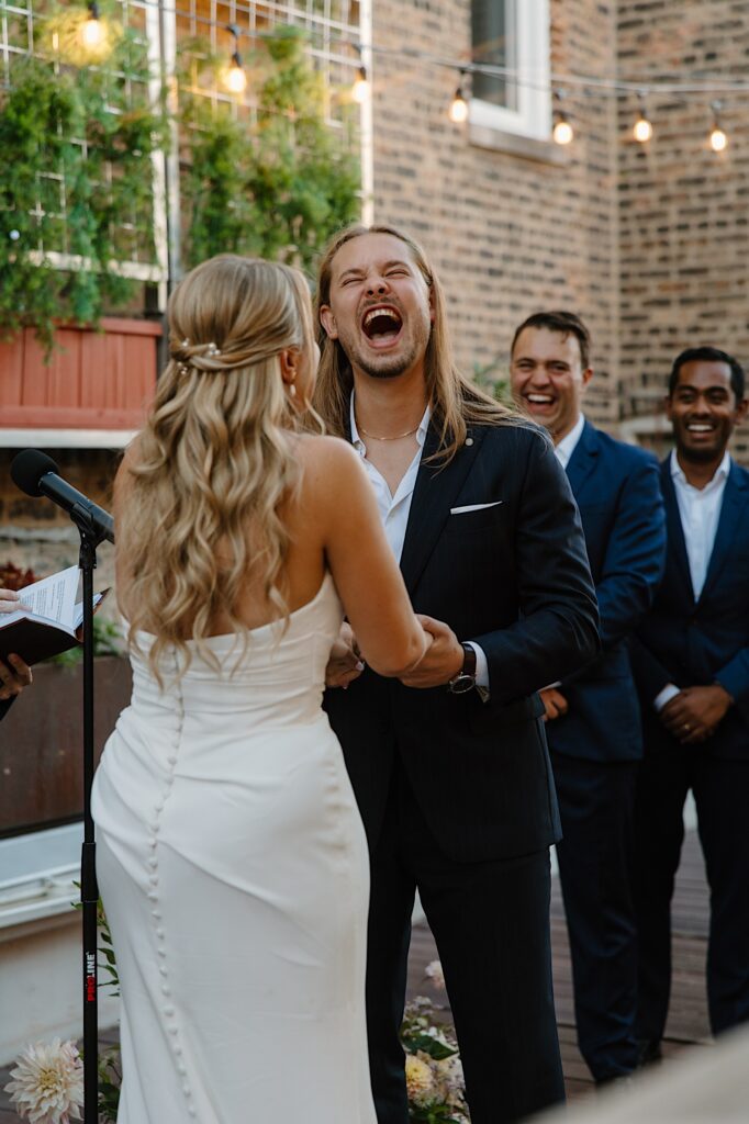 The groom laughs with his whole face at something his bride says during their wedding ceremony.  His groomsmen dressed in navy blue suits also laugh in the background.  