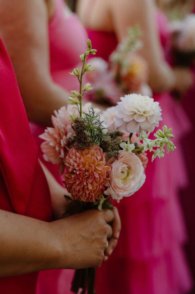 Bridesmaids stand in their hot pink bridesmaids dresses holding small bouquets of pink dahlias and ranunculus. 