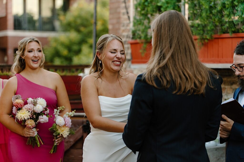 A bride laughs with her whole face while holding the hands of her groom during their Chicago rooftop wedding ceremony.  A bridesmaid stands behind her in a hot pink dress holding two bouquets of dahlias. 