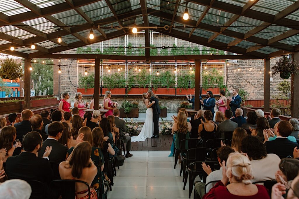 A bride and groom kiss concluding their rooftop wedding ceremony in Chicago.  Their guests sit under a covered structure and there are string lights hanging throughout the venue. 