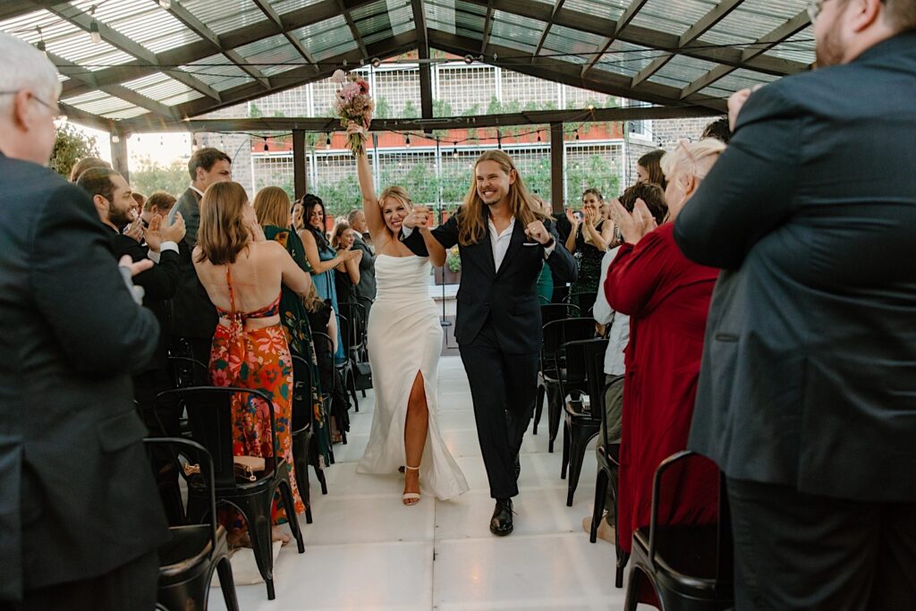 A bride and groom laugh and celebrate with guests as they walk down the aisle after their ceremony at their Chicago rooftop wedding venue Homestead on the Roof. 