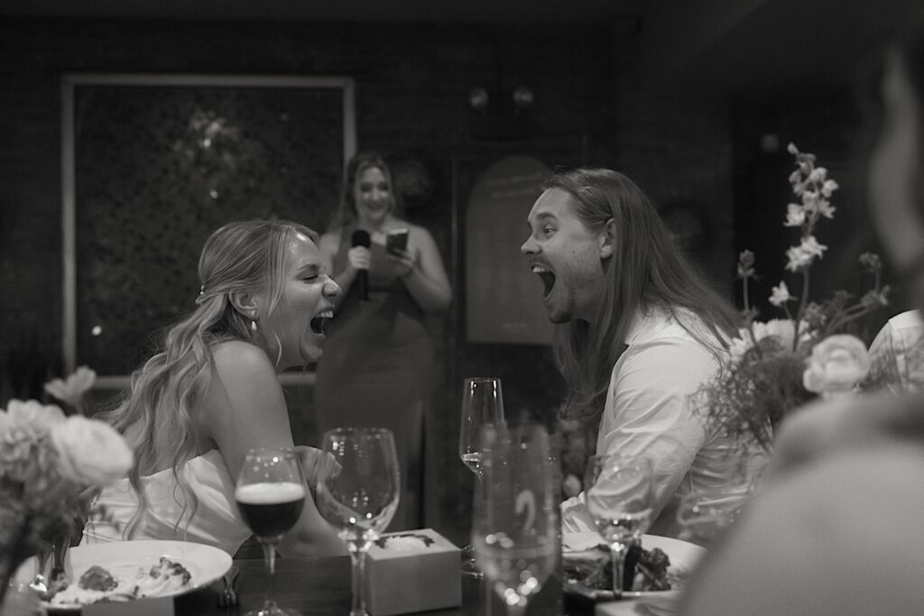 The bride and groom laugh together while a bridesmaid shares a story during her wedding toast.  Their reception is in the indoor portion of Homestead on the Roof. 