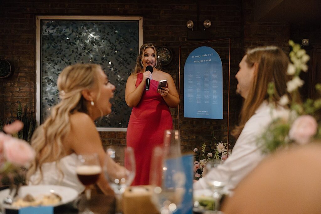 A bridesmaid in a hot pink wedding dress stands in front of a simple periwinkle blue seating chart sign as she gives a toast to the bride and groom who are in the foreground of the photo. 