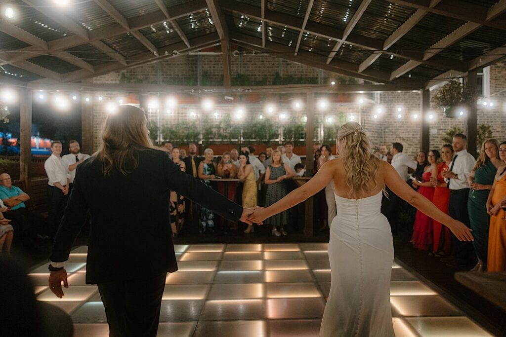 A bride and groom walk onto the lit dance floor that is lined with their guests for their first dance.  The rooftop wedding venue in Chicago is lit with string lights and the lighting within the dance floor. 