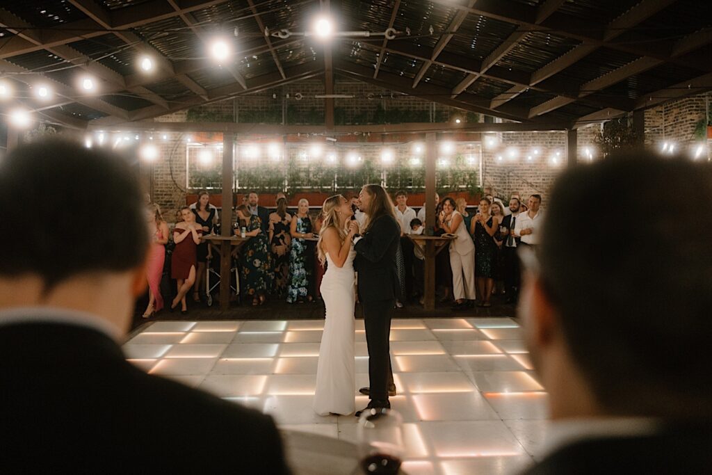 A bride and groom share a first dance on a lit dance floor that is lined with their guests for their first dance.  The rooftop wedding venue in Chicago is lit with string lights and the lighting within the dance floor. 