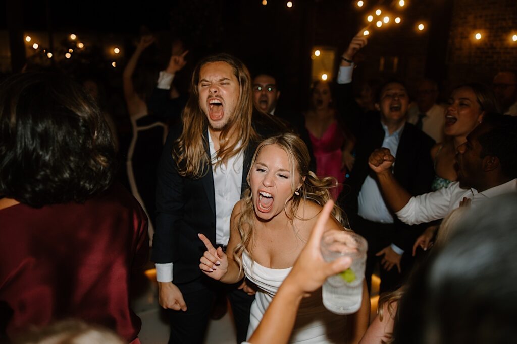 A bride and groom sing and dance together surrounded by guests at their Chicago rooftop wedding venue that is lit by string lights and lights in the dance floor. 