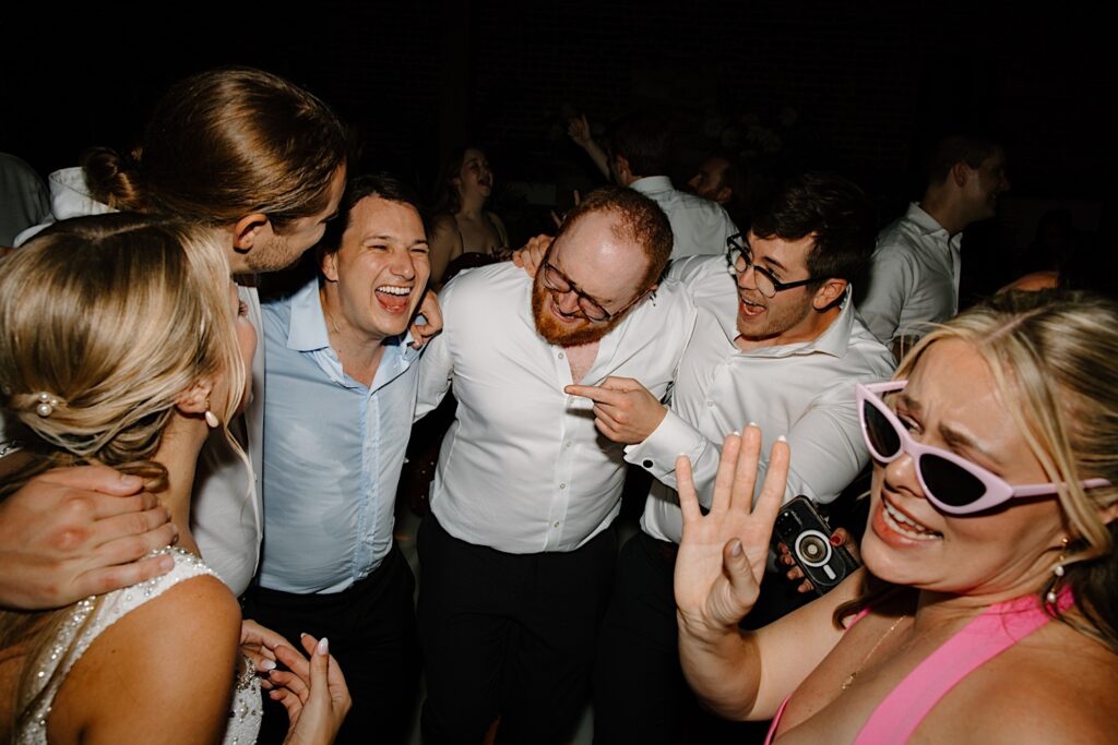 Wedding guests gather around the bride and groom to dance and sing at their wedding reception in Chicago.  A bridesmaid is in the foreground singing towards the camera with light pink sunglasses and a hot pink dress. 