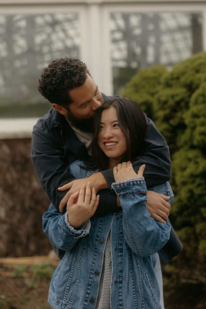 man kisses his wife's head with his arms around her while she smiles. 